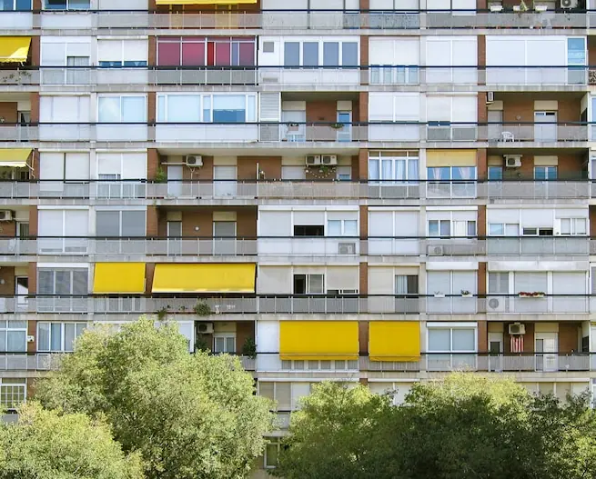 Rânduri de balcoane, unele cu copertine galbene, pe un bloc lung din spatele copacilor